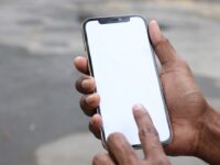 Main sombre tenant un smartphone doré à écran blanc vierge, un doigt effleurant la surface. Prêt pour l'affichage de noms d'IA.