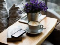Café latte, smartphone sur livre, fleurs violettes sur table en bois. Scène du quotidien évoquant l'intelligence personnelle et l'IA.