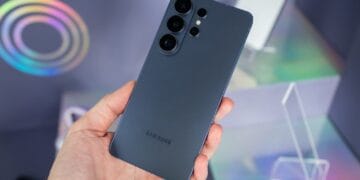 Main tenant un smartphone Samsung gris foncé, dos avec triple capteur photo, sur fond holographique. Illustre le Vibe Coding et le développement IA.