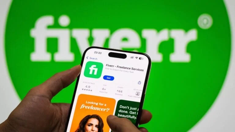 Main tenant un smartphone affichant l'application Fiverr pour services freelance, avec le logo vert Fiverr en arrière-plan. Idéal pour AI Logo by Fiverr.