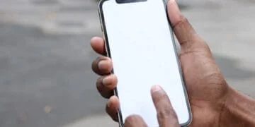 Main tenant un smartphone à cadre doré avec écran blanc, un doigt le touche. Évoque l'interaction avec Claude, Siri, Grok et les noms d'IA.