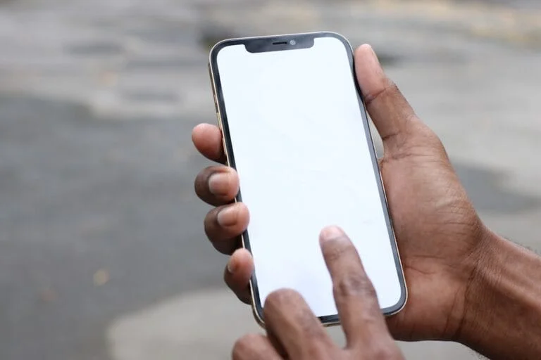 Main tenant un smartphone à cadre doré avec écran blanc, un doigt le touche. Évoque l'interaction avec Claude, Siri, Grok et les noms d'IA.