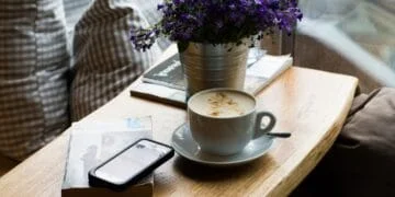 Tasse de café latte, smartphone et livre sur table en bois, fleurs violettes. L'IA s'invite dans votre quotidien.
