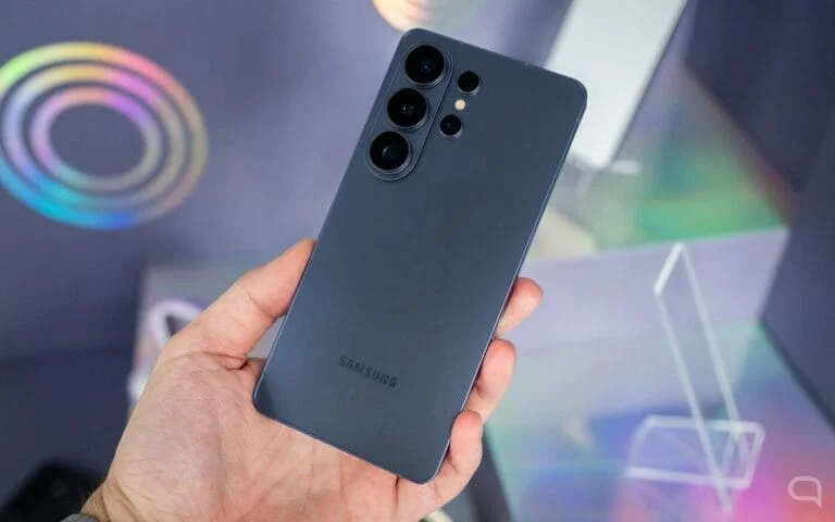 Hand holding a dark gray Samsung smartphone, back with triple photo sensor, on holographic background. Illustrates Vibe Coding and AI development.
